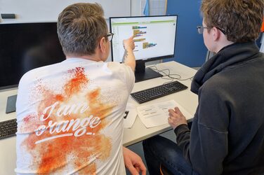 Zwei Personen sitzen an einem Tisch, eine von ihnen zeigt der anderen etwas auf einem Bildschirm.