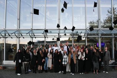 The graduates stand in front of the faculty building and throw their graduation hats in the air.