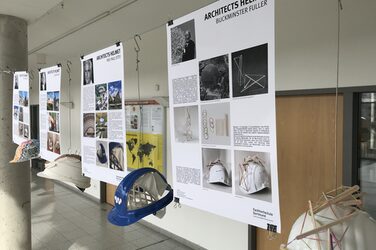 Das Bild zeigt vier erläuternde Plakate mit den dazugehörigen Architekten-Helmen, wie sie im Foyer des Fachbereichs Architektur hängen. Sie sind zum Beispiel in Anlehnung an Buckminster Fuller oder Frei Otto mit Holzstäbchen in besonderer Anordnung beklebt oder andersfarbig lackiert.