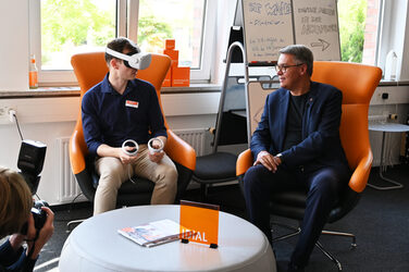 Jonas Sorgalla and Mayor Thomas Westphal talk about a VR application while sitting in wing chairs. Jonas is wearing VR glasses.