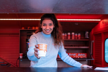 Eine Studentin reicht einen Kaffeebecher mit dem Slogan "Do you speak orange?"über den Tresen des Kaffeetrucks und lächelt dabei. A student hands a coffee cup with the slogan ‘Do you speak orange?’ over the counter of the coffee truck and smiles