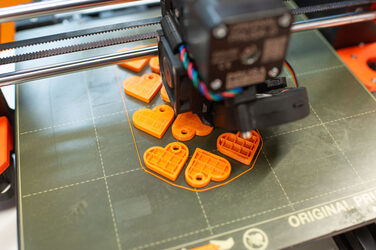 Ein 3D Drucker erstellt orangfarbene Herzen als Schlüsselanhänger. A 3D printer creates orange-colored hearts as key rings.