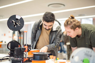 Ein Mitarbeit des FabLabs der Fachhochschule Dortmund erklärt einem interessierten Studierenden, wie der auf dem Tisch stehende 3D Druck funktioniert.An employee of the FabLab at Dortmund University of Applied Sciences and Arts explains to an interested student how the 3D printer on the table works.