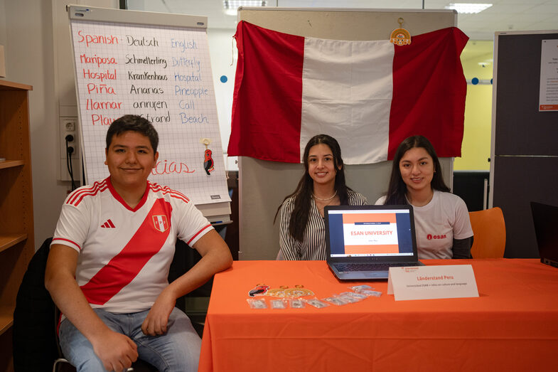 Drei Personen des Länderstand Peru sitzen an ihrem Stand und lächeln in die Kamera.__Three people from the Peru country stand are sitting at their stand and smiling at the camera.