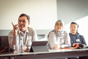 Drei Personen sitzen an einem Tisch und schauen interessiert nach links. Der Mann ganz links gestikuliert freundlich mit den Händen__Three people sit at a table and look to the left with interest. The man on the far left gestures kindly with his hands