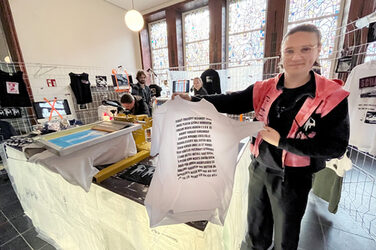 Ein Mensch in schwarzer Kleidung und roter Weste hält ein T-Shirt mit aufgedruckten Worten ins Bild. Er steht an einem Stand mit einer Siebdruckmaschine und weiteren Kleidungsstücken.