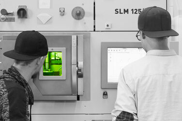 Two students look at a monitor and through a viewing window of a laser melting machine.