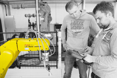 Two young men stand in front of an industrial robot in the robotics lab and control the machine.