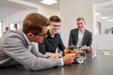 Foto von drei Personen an einem Tisch, auf dem ein 4-rädriger Roboter steht. Eine der Personen beugt sich so über den Tisch, dass er sich den hinteren Teil des Roboters angucken kann. Die anderen beiden Personen gucken aufmerksam zu und achten darauf, was die andere Person ihnen zeigen will.