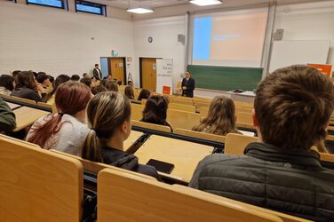 In einem Hörsaal sitzen Menschen in mehreren Reihen und schauen zu einer Person, die ganz vorne steht.