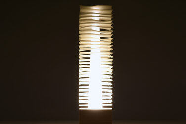 Night shot of a column-like, illuminating, sculptural luminaire on a wooden base