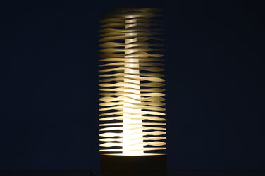 Night shot of a column-like, illuminating, sculptural luminaire on a wooden base