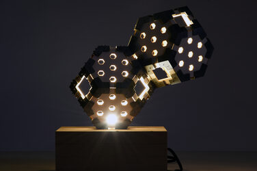 Night shot of an illuminated, sculptural hexagon luminaire on a wooden base with generatively manufactured elements