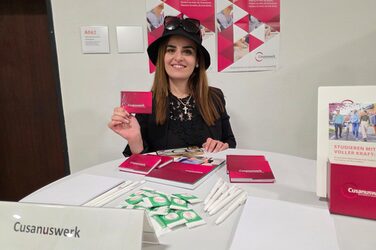 Eine Person mit Hut steht hinter einem Tisch und hält eine Karte mit der Aufschrift Cusanuswerk vor sich.