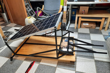 On the floor of a room is a wooden panel on which a metal frame with a solar module mounted at an angle stands.