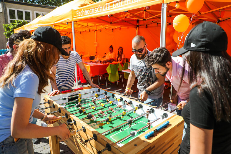 Mehrere Personen stehen auf einem Sommerfest um einen Kickertisch. Vier Personen kickern gegeneinander, zwei Personen schauen ihnen dabei zu.