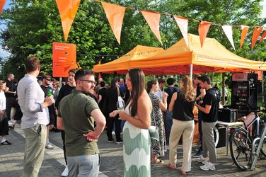 Menschen stehen auf dem Sommerfest des Fachbereichs Architektur der FH Dortmund. Unter orangefarbenen Pavillons versorgt die Fachschaft die Gäste mit Getränken.