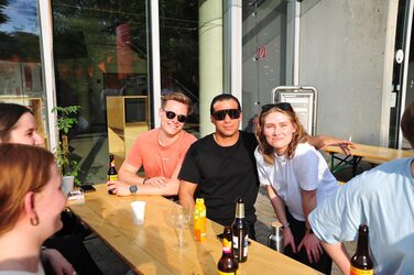 Gruppe junger Menschen sitzt entspannt bei Sonnenschein am Tisch vor dem Architekturgebäude – mit Getränken, Sonnenbrillen und guter Laune.