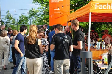 Menschen stehen auf dem Sommerfest des Fachbereichs Architektur der FH Dortmund. Unter orangefarbenen Pavillons versorgt die Fachschaft die Gäste mit Getränken.
