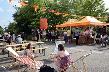Menschen sitzen und stehen auf dem Sommerfest des Fachbereichs Architektur der FH Dortmund. Unter orangefarbenen Pavillons versorgt die Fachschaft die Gäste mit Getränken.