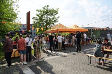 Menschen sitzen und stehen auf dem Sommerfest des Fachbereichs Architektur der FH Dortmund. Unter orangefarbenen Pavillons versorgt die Fachschaft die Gäste mit Getränken.