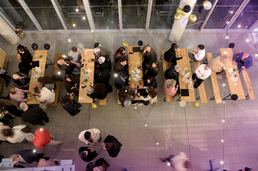 The guests sit and stand at long wooden tables in the faculty foyer, which is festively decorated with fairy lights and balloons. The atmosphere is convivial and relaxed.