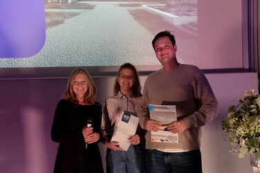 ChatGPT:

The dean and two students are standing on stage smiling into the camera, two of them holding certificates in their hands. A presentation slide and purple lighting can be seen in the background.
