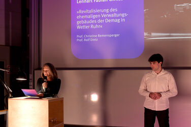 Dean Prof. Diana Reichle stands at the lectern and reads, while a student stands next to her on the stage. Behind them, the presentation slide shows a project with its title and name in purple light.