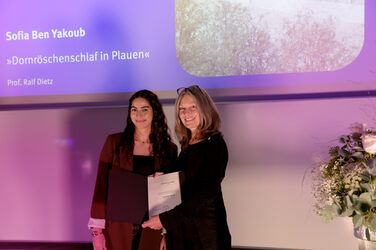The Dean congratulates a student on stage, they hold a certificate together, while a presentation slide with names and project title can be seen in the background. The scene is bathed in violet light.