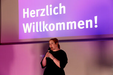Chancellor Svenja Stepper speaks with a microphone in front of a screen that reads "Welcome!" in large letters. The scene is bathed in violet light.