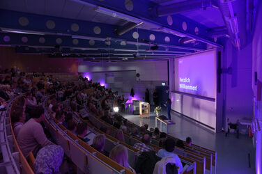 A packed lecture hall in which a woman is speaking at the front. The projection shows "Welcome!" in purple lighting.
