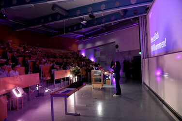 Presenter Eva Paar in the lecture hall in front of a large audience. The lighting is purple and "Welcome!" is written on the screen.
