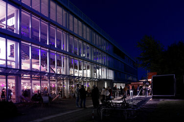 The faculty building is lit up in purple light in the evening, and people are celebrating inside and outside. In front of the building there are groups in conversation and a few deckchairs.