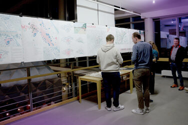Several people are looking at large-scale plans and models of urban development projects on the upper floor of an exhibition. The room has modern lighting and looks like part of a presentation or celebration.