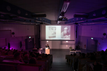 Dean Prof. Diana Reichle gives her lecture in a darkened lecture hall, while a book cover and a portrait are shown on the screen. The room is discreetly lit in violet.