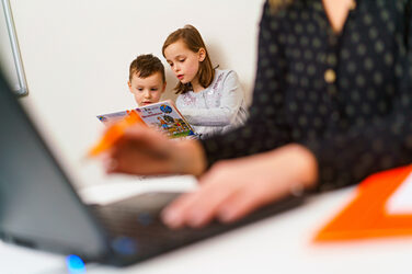 Foto von zwei jüngeren Kinder, die gemeinsam lesen. Im Vordergrund sieht man unscharf und im Anschnitt eine Person, die an einem Laptop arbeitet. __In the foreground you can see a laptop on the desk out of focus and someone who is working. The focus is on the children reading in the background.