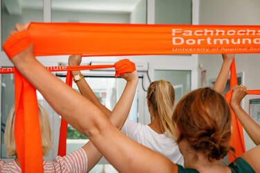 Foto mehrerer Personen, die mit orangenen Therabändern Pausensport machen.__ <br>Several people are doing break sports with orange thera bands.