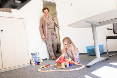 Foto eines kleinen Mädchens, das auf dem Boden eines Büros mit einer Holzeisenbahn spielt. Ihre Oma kommt zur Tür rein und lächelt auf sie herunter.__Photo of a little girl playing with a wooden train set on the floor of an office. Her grandma walks in the door and smiles down at her.