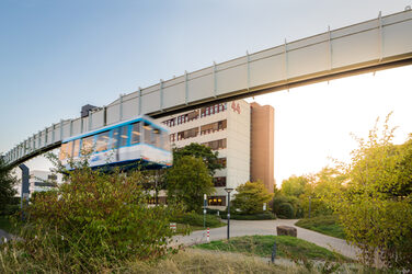 Foto der H-Bahn-Schienen mit vorbeifahrender H-Bahn, im Hintergrund des Gebäude Emil-Figge-Straße 44 der Fachhochschule Dortmund.