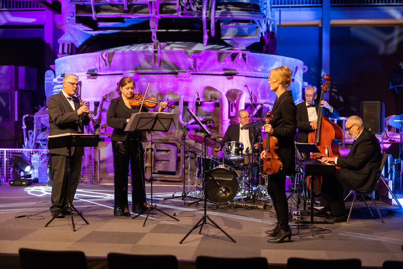 Foto einer Band, die auf einer Bühne spielt. Die Band besteht aus zwei weiblichen und vier männlichen Personen. Jede Person spielt ein Musikinstrument.