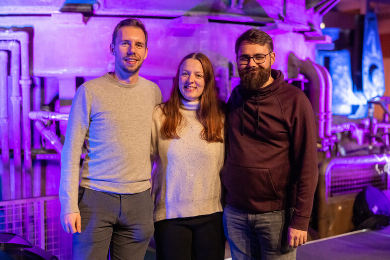 Foto von einer weiblichen und zwei männlichen Peronen die stehend in die Kamera lächeln.