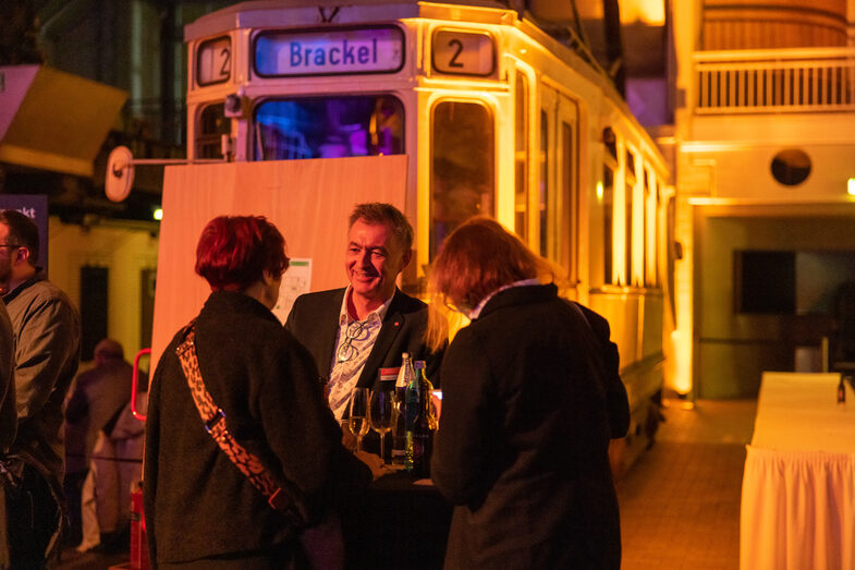 Foto einer männlichen und zwei weiblichen Personen. Diese stehen an einem Stehtisch vor einer alten Straßenbahn und unterhalten sich.