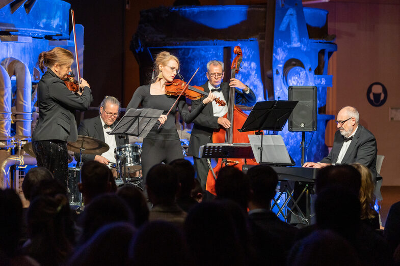 Foto einer Band, die auf einer Bühne vor einem Publikum spielt. Die Band besteht aus zwei weiblichen und drei männlichen Personen. Jede Person spielt ein Musikinstrument.