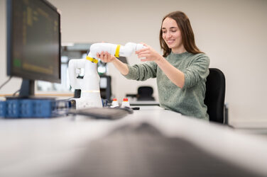 Foto einer Studierenden, die an einem Computertisch sitzt und mit der Hand den Arm eines Schrenkarmroboters hält.