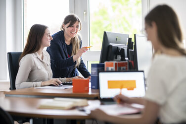 Foto von 3 Frauen an Schreibtischen. Links im Bild sitzt eine junge frau am Schreibtisch, von ihr links steht eine Frau neben ihr, die auf den Monitor zeigt. Rechts ist unscharf eine Frau mit Rücken zur Kamera, diese schreibt etwas