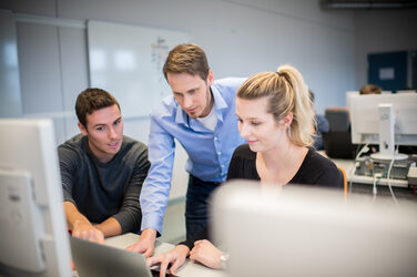 Foto eines Lehrenden im Computerraum, der zwischen einem Studenten und einer Studentin steht und beiden etwas am Laptop erklärt. __ <br>The professor stands between a male student and a female student in the computer room and explains something to both of them on the laptop.