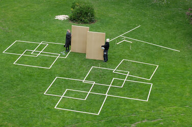 Students lay out the floor plan of their design on the green meadow in the inner courtyard of Fachhochschule Dortmund's Department of Design. They designed a maze that offers the opportunity to experience spatial perception in a new way. An inaccessible space forms the center of the design, which can only be glimpsed through mirrors and peepholes. The picture shows prefabricated rectangular frames made of roof battens, which are spread out on the lawn and reveal a preliminary arrangement. Students carry the first wall sections to the green area and discuss the next steps together.