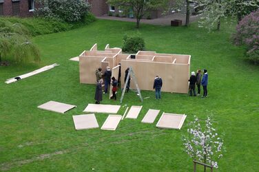 The floor plan is defined and some wall sections are attached so that the structures of a labyrinth are visible. The workshop students are spread out all around, surrounded by other components, roof battens and a ladder.