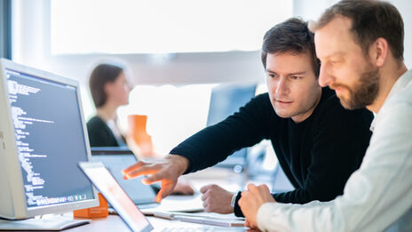 <br>Photo of two university IT employees looking at a laptop together at their workstation, one is pointing at something with his hand. Another employee is in the background.