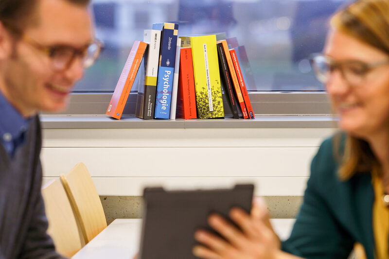 Foto von mehreren Bücher, die aneinander gelehnt auf einer Fensterbank stehen. Im Vordergrund zwei unscharf zu erkennende Personen, die sich austauschen.